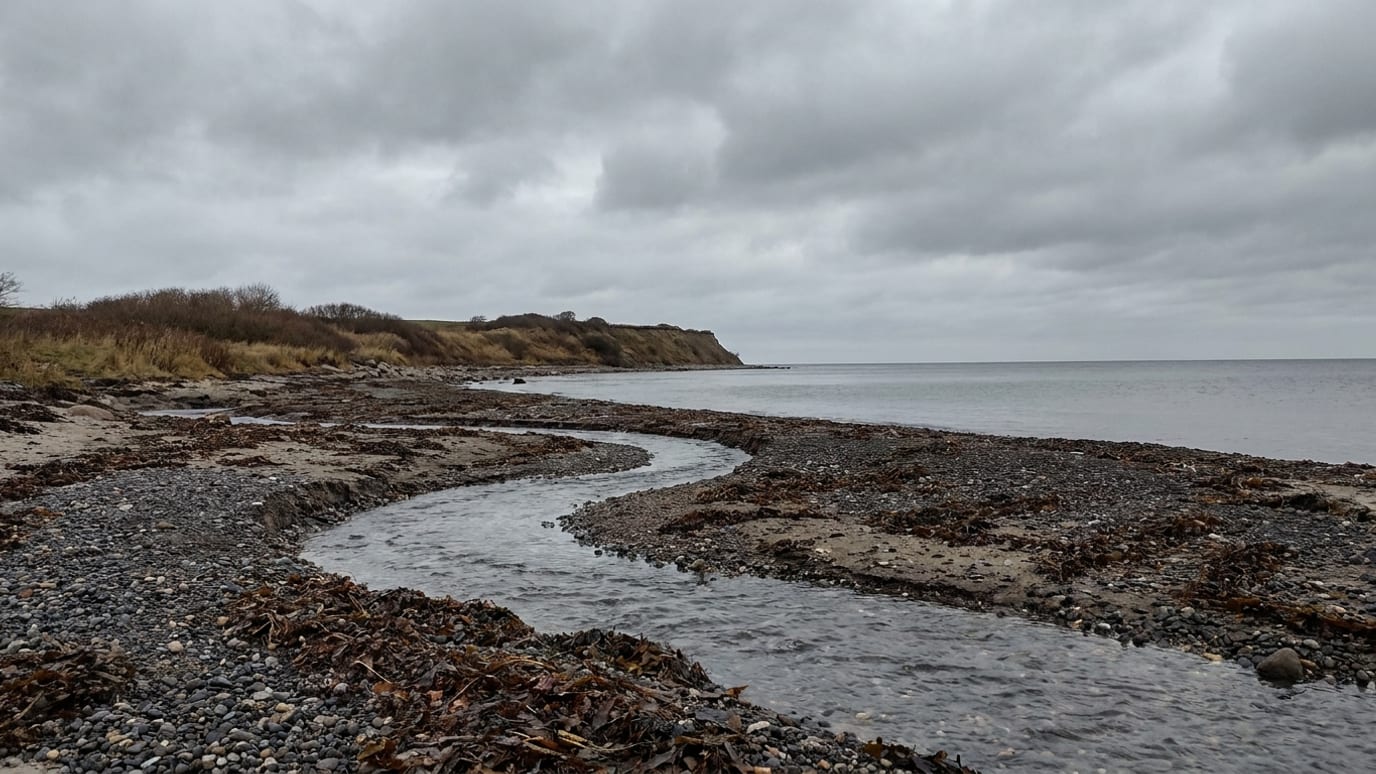 Et lille vandløb løber ud over en stenstrand og ind i havet en grå efterårsdag